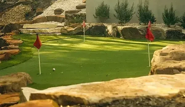 Golf area with putting green, three red flags, and rocks surround the border with a white house with blue accents in the background.