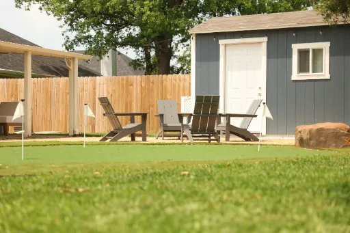Residential backyard putting green with white flags, gray shed, Adirondack chairs, and wooden privacy fence.