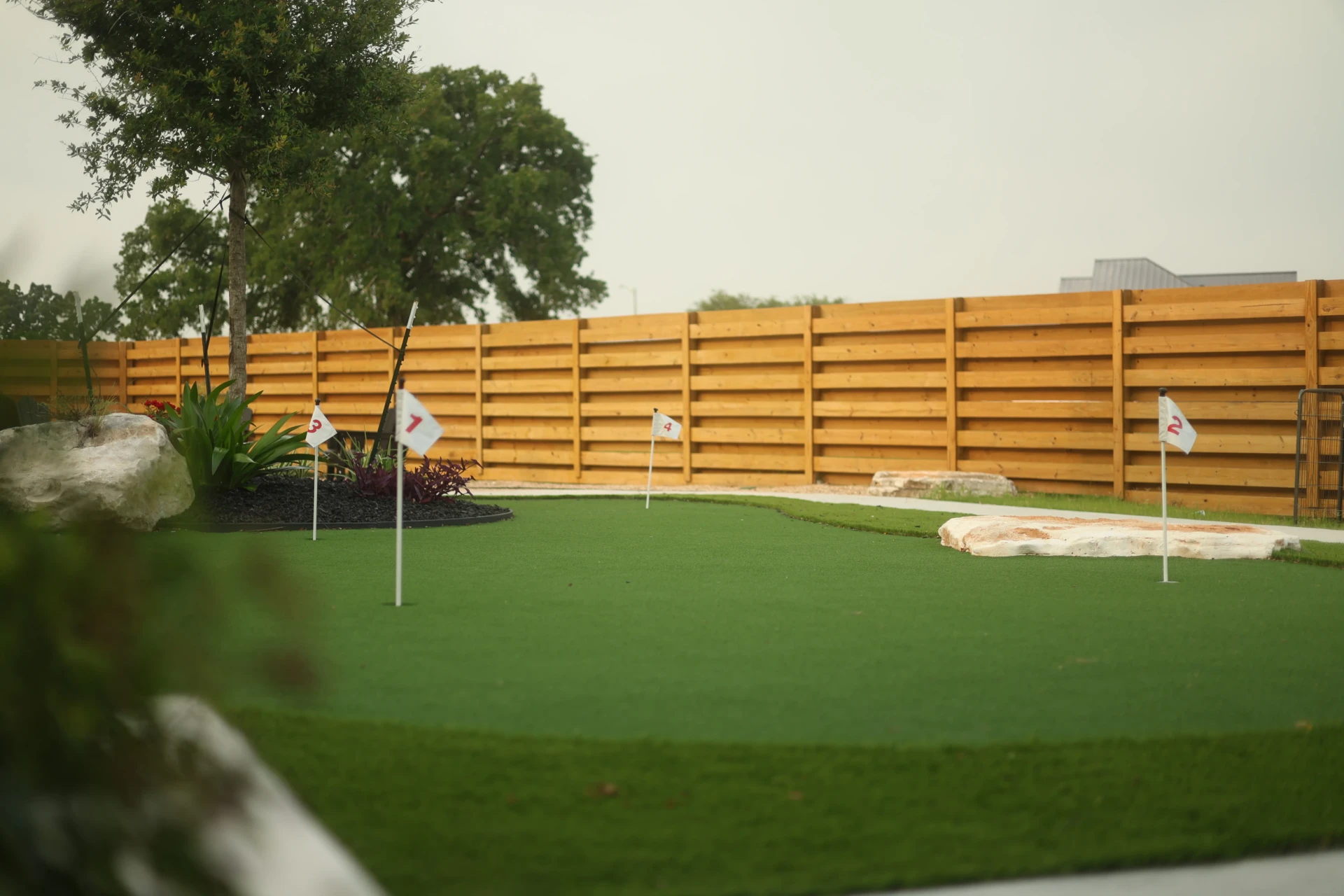 Large backyard putting green with multiple white flag holes, wooden fence, and landscaped trees and plants.