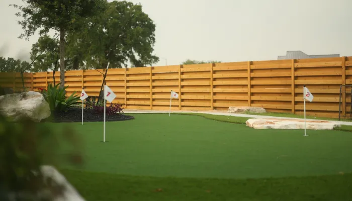 Large backyard putting green with multiple white flag holes, wooden fence, and landscaped trees and plants.