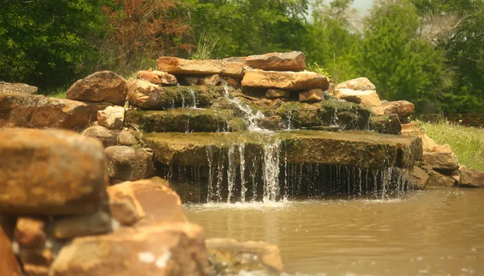Natural stone waterfall feature flowing into a backyard pond surrounded by greenery.