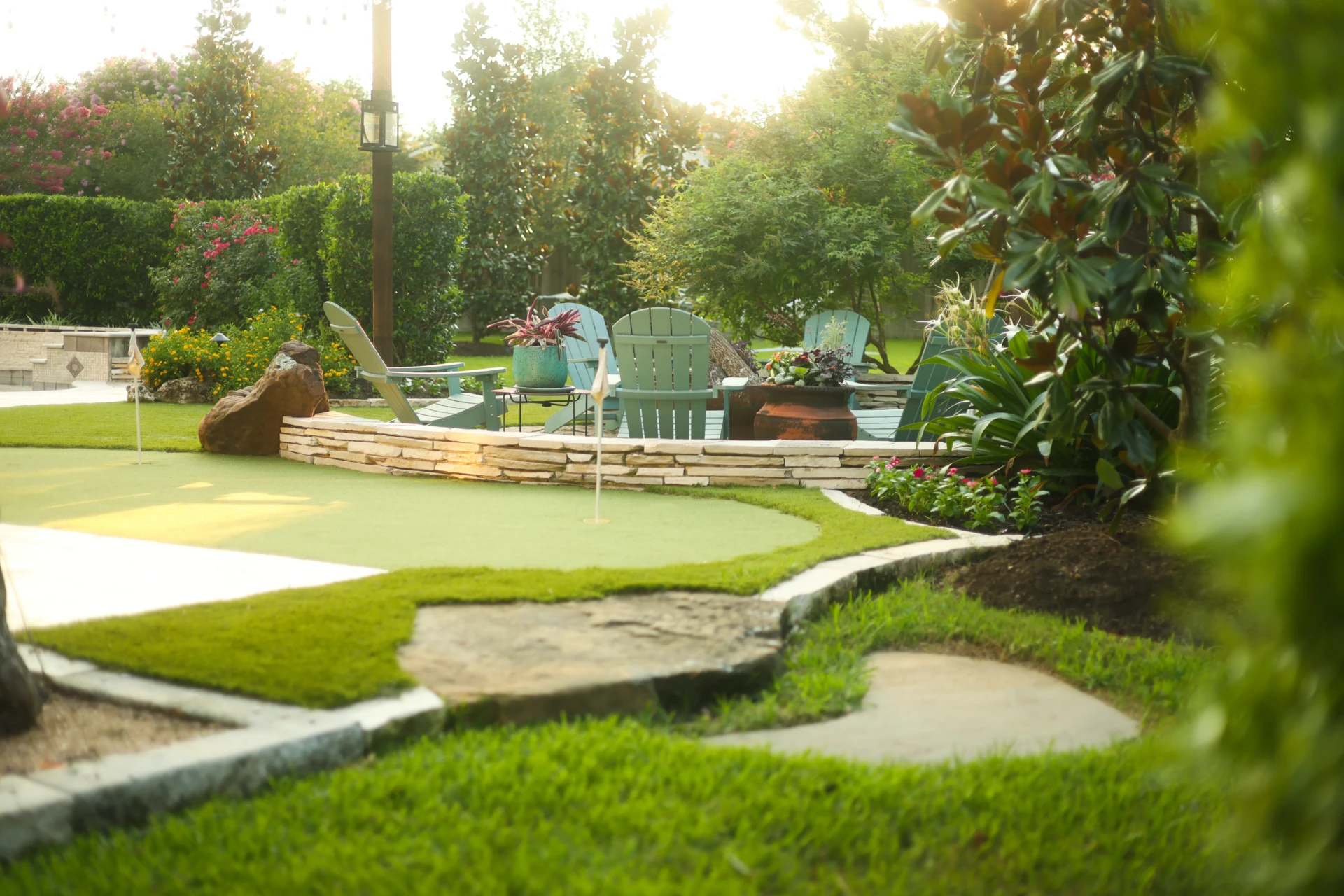 Backyard putting green next to stone fire pit and Adirondack chairs surrounded by colorful plants and greenery.