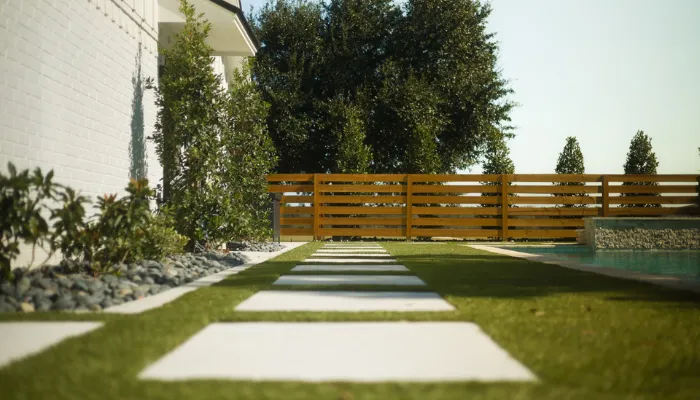 Stepping-stone walkway set into artificial turf beside a modern home, with clean landscape edging, low plantings, river rock borders, and a wood privacy fence near a backyard pool.